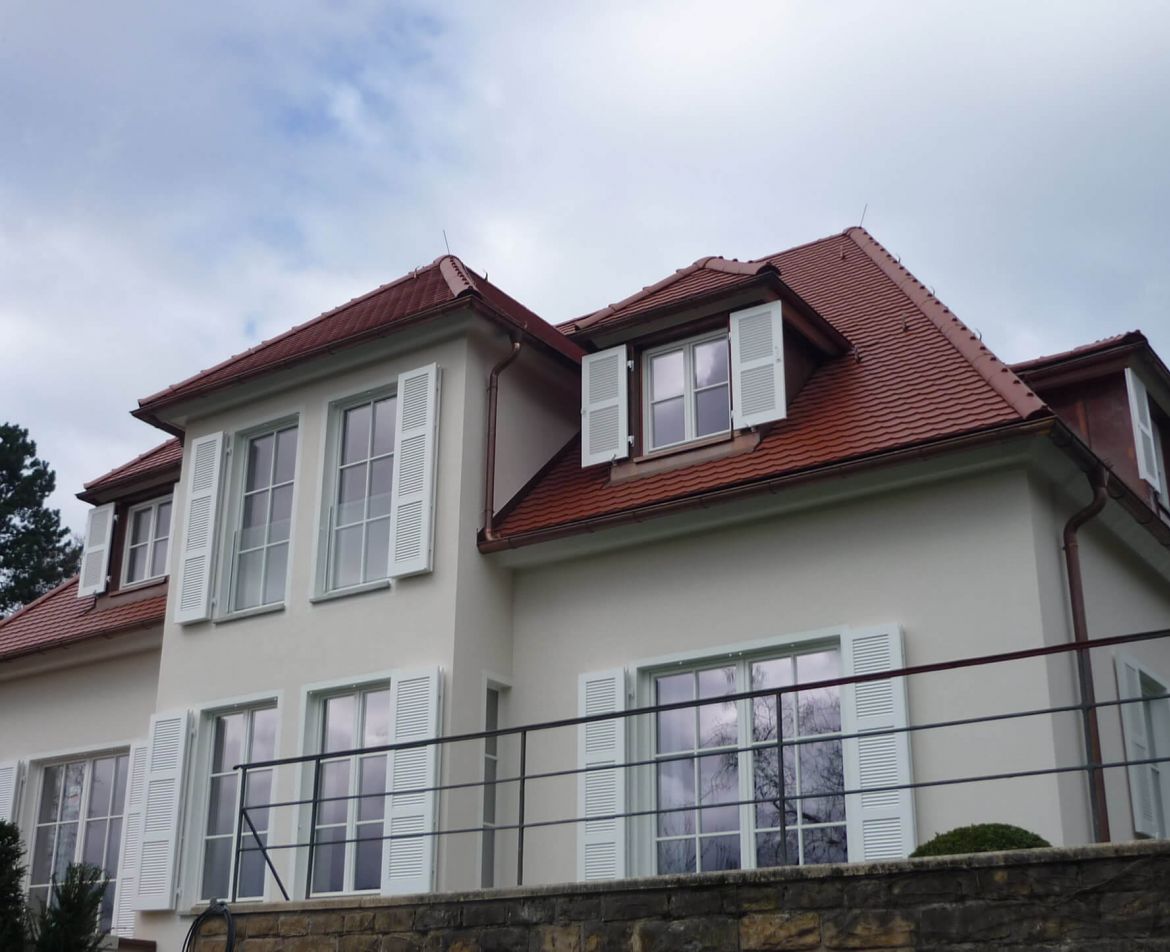 Renovierte Altbau-Fassade mit rotem Ziegeldach, wei&szlig;en Fensterl&auml;den und einem umlaufenden Balkon